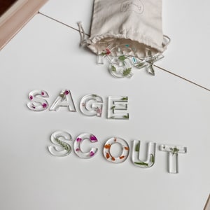 May include: Clear resin letters spelling out "SAGE SCOUT" with pressed flowers and greenery inside. The letters are on a white surface and a small white bag with the text "leaf & branch" is in the background.