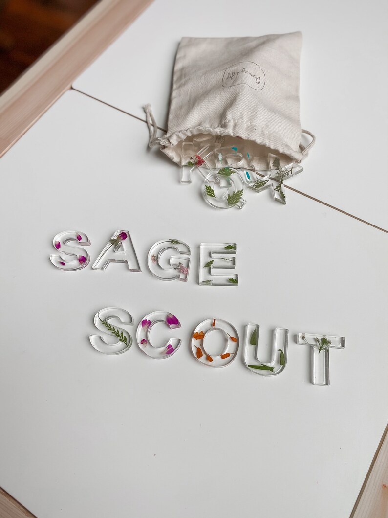 May include: Clear resin letters spelling out "SAGE SCOUT" with pressed flowers and greenery inside. The letters are on a white surface and a small white bag with the text "leaf & branch" is in the background.