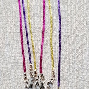 Könnte beinhalten: Fünf Paar Brillenketten mit durchsichtigen Plastikenden und silbernen Metallverschlüssen. Die Ketten sind aus Satinband in Lila, Rosa und Gold.