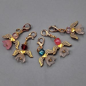 May include: Four gold-colored angel charms with wings, each featuring a different colored bead and a flower. The charms have lobster claw clasps for easy attachment. The beads are pink, red, blue, and purple, and the flowers are pink and white.