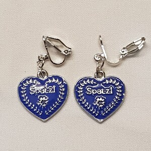 May include: A pair of blue heart-shaped earrings with a silver clip-on fastening. The hearts feature a white floral design and the word "Spaetzle" written in silver.