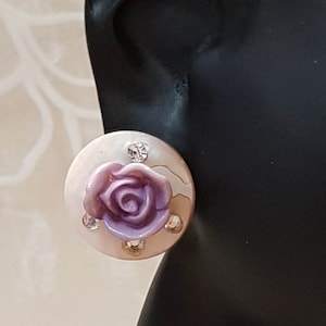 Puede incluir: Un pendiente redondo de color crema con un diseño de rosa púrpura en relieve en el centro. El pendiente está adornado con pequeños y brillantes pedrería. El pendiente está sobre un fondo negro.