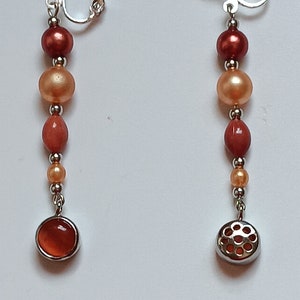 Könnte beinhalten: Ein Paar Ohrclips mit einer herabhängenden Perlenkette. Die Perlen sind orange, pfirsichfarben und braun. Die unterste Perle ist eine runde silberfarbene Perle mit einem geometrischen Muster.