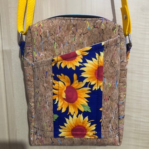 May include: A small cork and fabric crossbody bag with a yellow strap. The bag has a blue fabric pocket with a sunflower pattern. The cork fabric is brown with multi-coloured flecks.