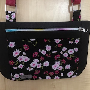 May include: Black crossbody bag with a zip closure and a floral print lining. The bag has a pink strap and a label that says "Handmade with love".