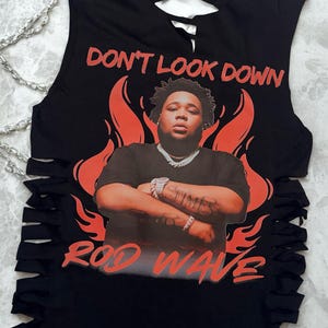 May include: A black sleeveless shirt with side cutouts and a crisscross neckline. The graphic features a portrait of Rod Wave surrounded by orange flames, with the text DON'T LOOK DOWN at the top and ROD WAVE at the bottom in orange lettering.