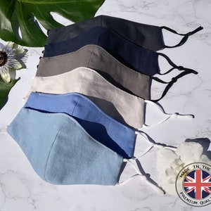 May include: Five face masks in different colours, including light blue, grey, beige, dark blue, and black. The masks are made of fabric and have adjustable ear loops. The masks are arranged in a stack, with the light blue mask on top. The masks are on a white marble surface. A green leaf and a white flower are in the background. The masks have a label that says "Made in the UK Premium Quality".