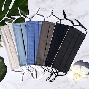 Può includere: Una collezione di mascherine plissettate in vari colori, tra cui bianco sporco, azzurro, denim, grigio, blu navy e nero. Ogni mascherina ha elastici regolabili bianchi o neri. Le mascherine sono disposte su una superficie di marmo bianco, con foglie e fiori tropicali sullo sfondo.