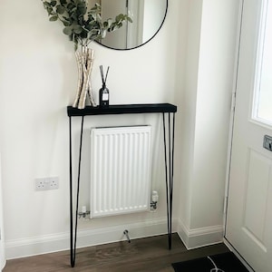 May include: A black console table with a thin black top and three hairpin legs. The table is decorated with a vase of dried branches and a black diffuser bottle. The table is placed in front of a white radiator.