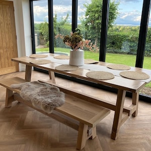 May include: A long wooden dining table with a natural finish and a bench seat. The table has a white vase with a bouquet of flowers on it. The table is set with placemats and is in front of a large window with a view of a green lawn.