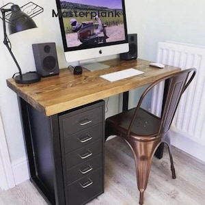 May include: A rustic wooden desk with black metal legs and a black metal filing cabinet with five drawers. A copper metal chair is positioned to the right of the desk. A computer monitor with a screen displaying a photo of a scooter is on the desk. A black lamp with a black shade is on the left side of the desk. Two black speakers are on the desk.
