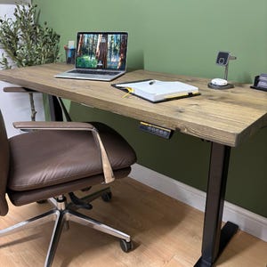 May include: A wooden standing desk with a laptop, notebook, and phone charger. The desk has a brown, textured surface and black legs. A brown leather office chair is in front of the desk. The laptop screen displays a forest scene.
