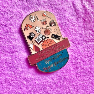 May include: A gold-toned enamel pin with a pink and blue background. The pin features a brain with a variety of colorful icons, including a paintbrush, a book, a musical note, and a tent. The text "Attention Deficit Hyperactivity Disorder" is written in red and white on a pink background. The text "Operation Hyperfixation" is written in white on a blue background.