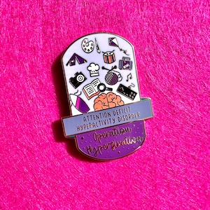 May include: A gold-coloured enamel pin with a purple and blue background. The pin features a brain with a thought bubble filled with various icons, including a paintbrush, a tent, a book, and a musical note. The text on the pin reads "Attention Deficit Hyperactivity Disorder Operation Hyperfixation."