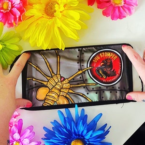 May include: A black and green face mask with a graphic of a bug-like creature with the text "Bug Stomper" and "We Endanger Species" in a red circle.