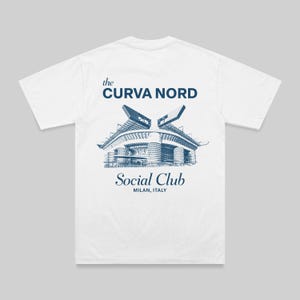 May include: White t-shirt with a blue graphic of a stadium and text that reads "the CURVA NORD Social Club MILAN, ITALY". The design is a detailed illustration of a building. The shirt has short sleeves and a crew neck.