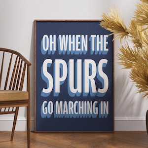 May include: A framed print with the text "OH WHEN THE SPURS GO MARCHING IN" in white and blue lettering on a navy blue background. The print is in a brown wooden frame and is displayed near a wooden chair and decorative plant.