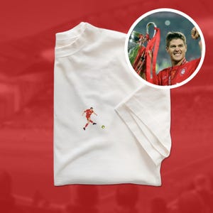 May include: White t-shirt featuring a small graphic of a soccer player kicking a ball. A circular image of a soccer player holding a trophy is in the upper right corner. The shirt is on a red background.