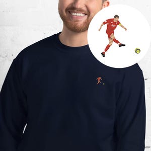 May include: Navy blue sweatshirt with a small embroidered soccer player graphic on the chest. A larger circular graphic shows a soccer player kicking a ball. The player is wearing a red uniform.