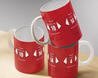 May include: Red ceramic mugs with white handles and interiors. Each mug features a graphic of a soccer jersey with a player's name and number. The mugs are stacked on a light brown surface.