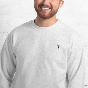 Pode incluir: Uma camisa de moletom cinza com uma pequena imagem bordada de um &aacute;rbitro levantando a m&atilde;o no ar.