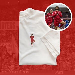 Puede incluir: Camiseta blanca con una imagen roja de un jugador de fútbol en una pose de yoga. El jugador lleva una camiseta roja y pantalones cortos blancos. La imagen está centrada en la camiseta. El fondo es una imagen borrosa de un estadio de fútbol.