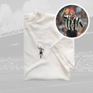 May include: White t-shirt with a black and white embroidered image of a soccer player celebrating a goal. The player is wearing a black and white striped jersey with the number 9 on the back. The t-shirt is folded and laid flat on a surface.