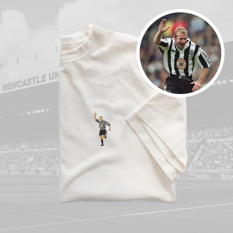 May include: White t-shirt with a black and white embroidered image of a soccer player celebrating a goal. The player is wearing a black and white striped jersey with the number 9 on the back. The t-shirt is folded and laid flat on a surface.