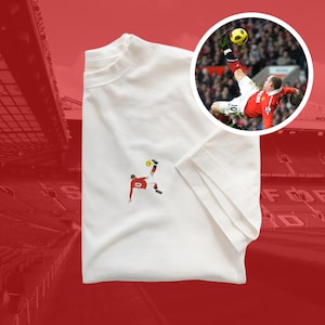 Puede incluir: Camiseta blanca con una pequeña imagen bordada de un jugador de fútbol pateando un balón. El jugador lleva un uniforme rojo y blanco. El fondo es un estadio rojo.