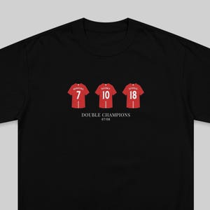 May include: Black t-shirt featuring three red soccer jerseys with player names and numbers: Ronaldo 7, Rooney 10, and Scholes 18. Below the jerseys, the text reads "DOUBLE CHAMPIONS 07/08".