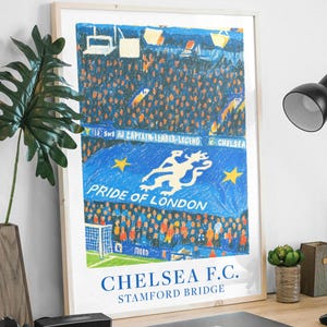 May include: A framed art print of a soccer stadium scene. The artwork features a blue banner with a white lion and the text "Pride of London." The words "Chelsea F.C. Stamford Bridge" are at the bottom. The print is displayed on a wooden desk.