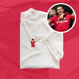 Könnte beinhalten: Weißes T-Shirt mit einer roten Comicfigur eines Fußballspielers, der einen Treffer mit erhobenen Armen feiert. Das T-Shirt hat ein kleines rotes gesticktes Logo der Social-Media-Plattform TikTok.