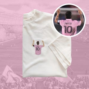 May include: White t-shirt featuring an embroidered image of a football player wearing a pink jersey with the number 10 and the name "Messi" on the back.