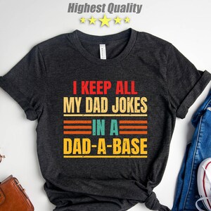 May include: Dark gray t-shirt with the text "I KEEP ALL MY DAD JOKES IN A DAD-A-BASE" in red, orange, and yellow. The shirt is styled with blue jeans and white sneakers.