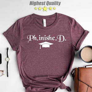 May include: A maroon t-shirt with the text "Ph.inished.D." in white with a graduation cap graphic. The shirt is laid flat on a white surface with other accessories.