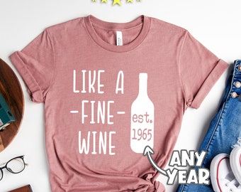 May include: A pink t-shirt with the text "Like a -Fine- Wine est. 1965 Any Year" printed on it. The shirt is laid flat on a white surface with a wooden cutting board, a pair of glasses, a brown wallet, a camera, and a pair of white sneakers.