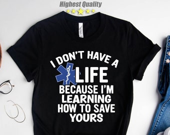 May include: Black t-shirt with a white graphic that says "I don't have a life because I'm learning how to save yours." The graphic also includes a blue and white medical star of life symbol.