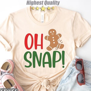 May include: A beige t-shirt with the text "OH SNAP!" in red and green letters. A gingerbread man cookie is falling apart, with brown icing and red candy details. The shirt is laid out on a white surface with a pair of pink Converse shoes and a pair of brown sunglasses.