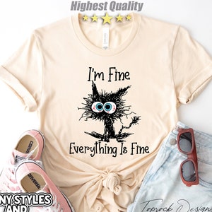 May include: A beige t-shirt with a black cat illustration and the text "I'm Fine Everything Is Fine".