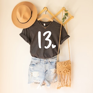 May include: A dark gray t-shirt with a white graphic of the number 13 and a small heart. The shirt is hanging on a wooden hanger with a straw hat and a woven bag.