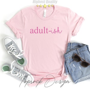 May include: Pink t-shirt with the text "adult-ish" in pink lettering. The shirt is paired with denim shorts and gray sneakers.