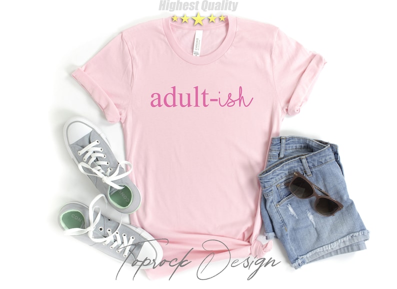 May include: Pink t-shirt with the text "adult-ish" in pink lettering. The shirt is paired with denim shorts and gray sneakers.