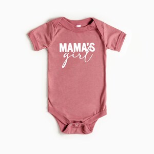 May include: A dusty rose baby bodysuit with the words "MAMA'S girl" printed in white. The short-sleeved onesie has a round neckline and snap closures at the bottom.