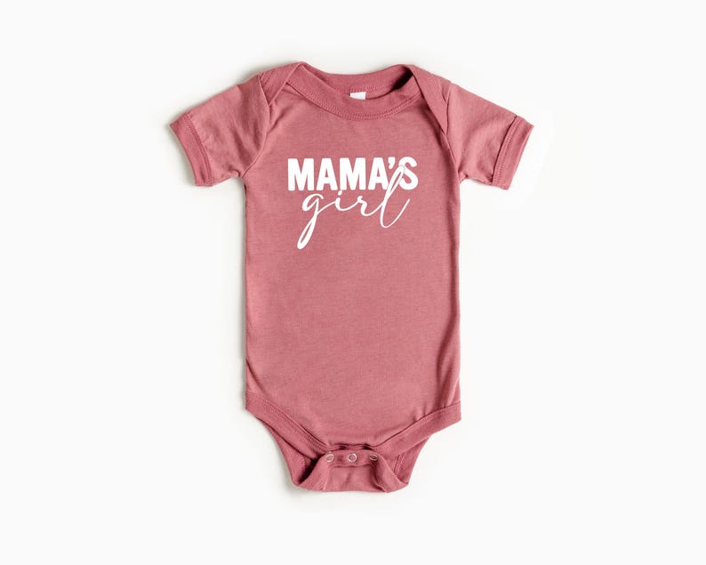 May include: A dusty rose baby bodysuit with the words "MAMA'S girl" printed in white. The short-sleeved onesie has a round neckline and snap closures at the bottom.