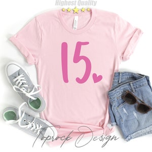 May include: Pink t-shirt with the number 15 in pink and a small pink heart. The shirt is on a white background with a pair of gray sneakers and denim shorts.