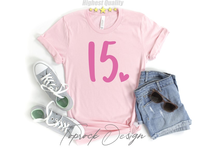 May include: Pink t-shirt with the number 15 in pink and a small pink heart. The shirt is on a white background with a pair of gray sneakers and denim shorts.