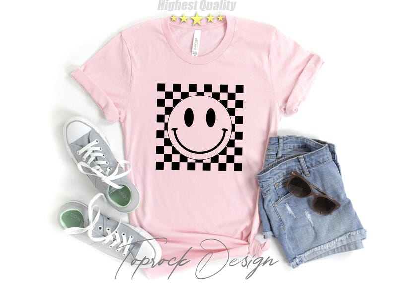 May include: Pink t-shirt with a black and white checkered pattern and a black smiley face in the center. The text "Toprock Design" is printed on the bottom of the shirt.