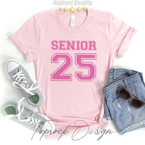 May include: Pink t-shirt with the text "SENIOR 25" in a pink athletic font. The shirt is laid flat on a white surface with a pair of gray sneakers and denim shorts.