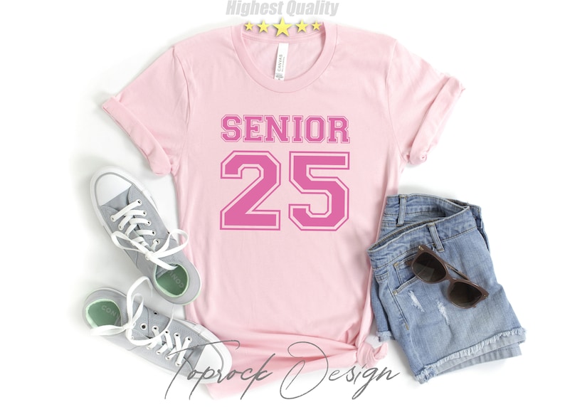 May include: Pink t-shirt with the text "SENIOR 25" in a pink athletic font. The shirt is laid flat on a white surface with a pair of gray sneakers and denim shorts.