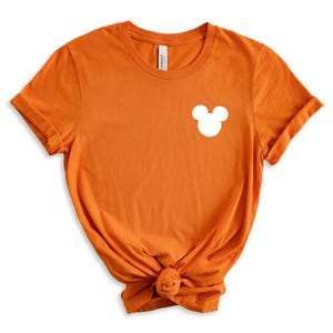 Puede incluir: Camiseta naranja de manga corta con una silueta blanca de Mickey Mouse en el pecho izquierdo.